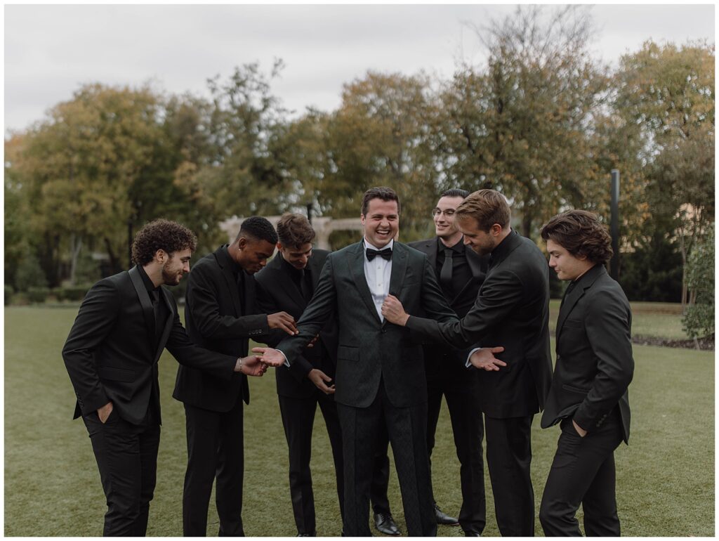 Groomsmen wearing all black making adjustments to groom's attire while groom is laughing 