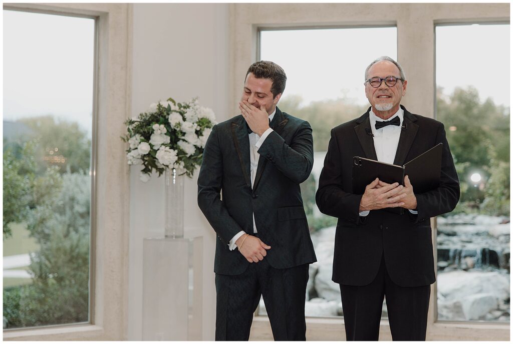 Groom standing at altar with officiant during wedding ceremony, smiling and covering his mouth. 