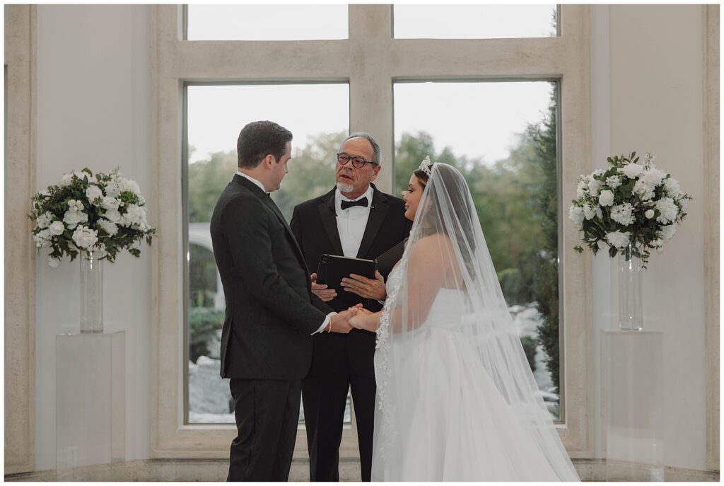 Bride and groom holding hands and standing in front of officiant during wedding ceremony. 