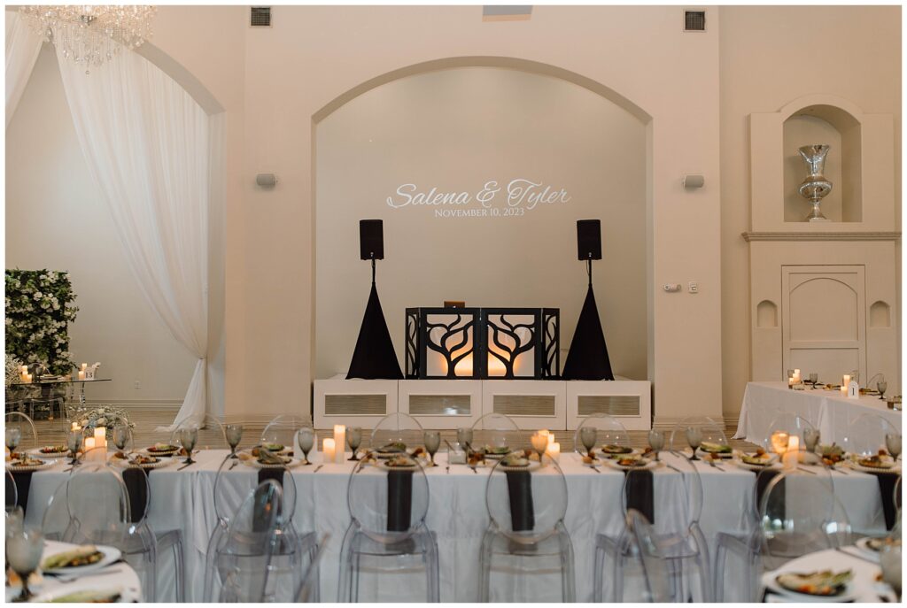 Reception space at Knotting Hill Place set with clear acrylic chairs, white table cloths with black accents. DJ stand is in the background with lights that say "Salena & Tyler" 