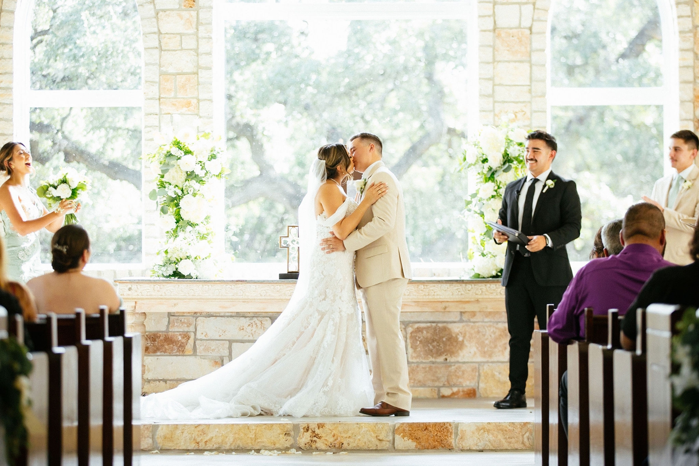 Bride and groom first kiss during wedding ceremony in a chapel with a stone wall and large windows