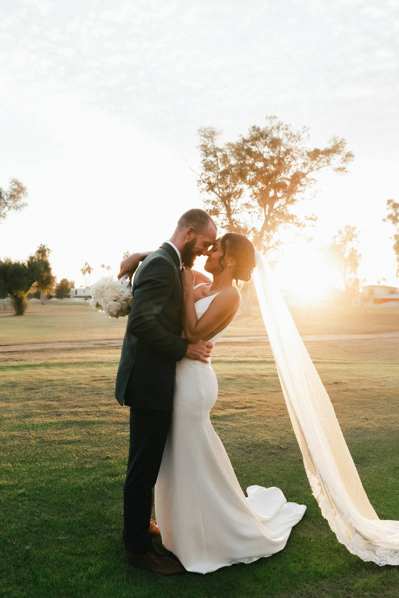 Bride embracing her groom with her veil flowing behind her at sunset
